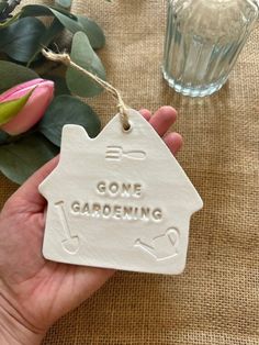 a hand holding a ceramic ornament that says gone gardening in front of some flowers