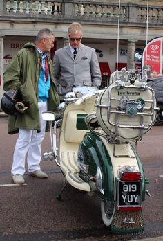 two men standing next to an old fashioned motor scooter on the street in front of a building