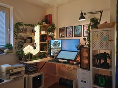 a desk with two computers on it in front of a window and some shelves filled with plants