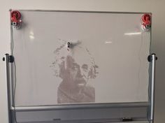 a white board with some wires attached to it and a drawing on the wall behind it