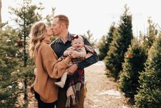 a man and woman kissing while holding a baby in front of christmas trees at the tree farm