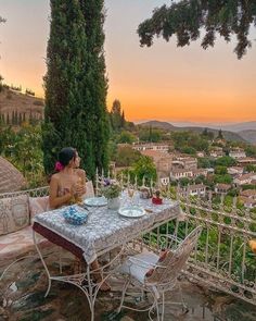 a woman sitting at a table on top of a cliff overlooking a town and trees