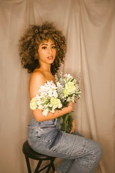 a woman is sitting on a stool with flowers in her lap and she has curly hair