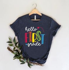 a t - shirt with the words hello first grade on it next to a plant