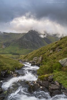 a stream running through a lush green valley under a cloudy sky with mountains in the background