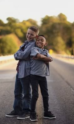 two boys hugging each other while standing on the side of an empty road with trees in the background