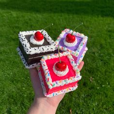 a hand holding three cakes with cherries on them in front of the camera and grass