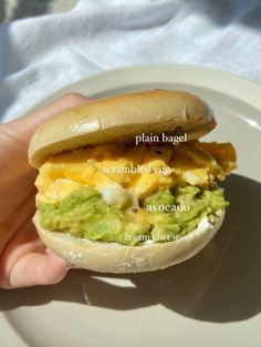 a hand holding a sandwich on a white plate with the words plan bagel scrambler egg avocado