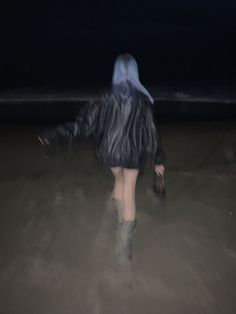 a woman in boots walking on the beach at night with her coat over her head