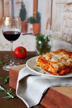 a white plate topped with lasagna next to a glass of wine and tomatoes