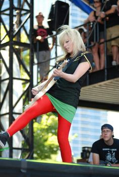 a woman in red and green tights playing guitar on stage with other people watching