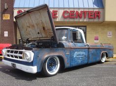 an old pickup truck with its hood open in front of a storefront and parking lot