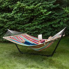 a woman laying in a colorful hammock reading a book on the grass next to some trees