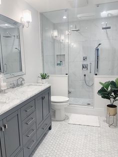 a white bathroom with gray cabinets and marble counter tops is pictured in this image, there are two plants on the floor next to the shower