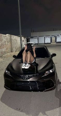 a woman sitting on the hood of a black sports car while holding a cell phone