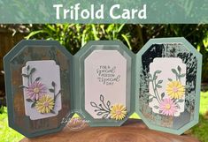 three cards with flowers on them sitting on top of a piece of wood in the grass