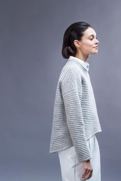 a woman standing in front of a gray background wearing a sweater and white pants with her hands on her hips