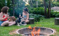 two women sitting next to a fire pit with dogs drinking wine in front of them