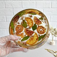 a hand holding a glass dish with orange slices and leaves on it, next to gold utensils