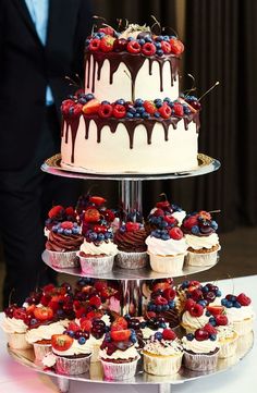 a three tiered cake with berries and chocolate on the top is surrounded by cupcakes