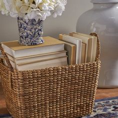 a wicker basket with books and flowers in it sitting on a rug next to a vase