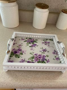 a white tray with purple flowers on it sitting on a counter next to three jars