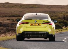 the rear end of a yellow bmw car driving down a country road with hills in the background