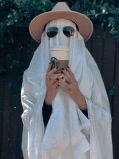 a person in a ghost costume holding a cup of coffee and wearing sunglasses with their face covered by a white cloth