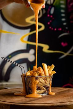 a person pouring sauce over french fries in a basket on top of a wooden table