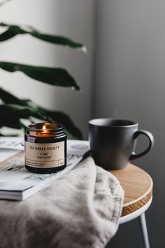 a candle sitting on top of a wooden table next to a cup and plant in the background