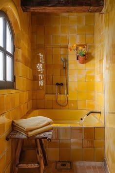 a yellow tiled bathroom with a window and bathtub in the corner, next to a wooden stool