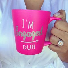 a woman holding a pink coffee cup with the words i'm engaged duh on it