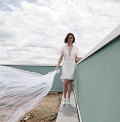 a woman is standing on a ledge with a veil in her hand and she is wearing a white dress