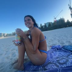 a woman is sitting on the beach holding a drink and smiling at the camera, with buildings in the background