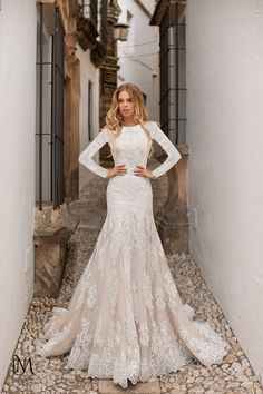 a woman is standing in an alley way wearing a long sleeved wedding dress with sheer lace