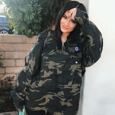 a woman in camo jacket leaning against a wall with her hand on her head