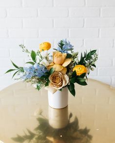 a white vase with yellow and blue flowers on top of a gold table next to a brick wall