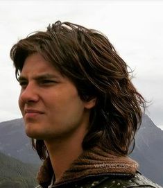 a young man with long hair standing in front of mountains and looking off into the distance