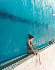 a woman sitting on the edge of a wall next to a swimming pool with blue water