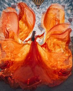 a woman in an orange dress is standing with her arms spread out and there is a hot air balloon flying overhead