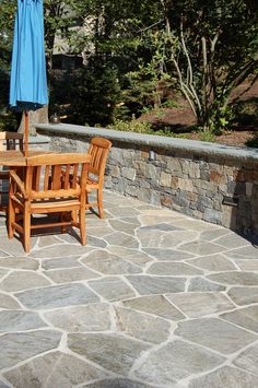 a wooden table and chairs sitting on a stone patio with an umbrella in the background