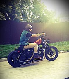 a man riding on the back of a motorcycle down a street next to a lush green field