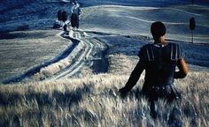 two men are walking in the middle of a field with tall grass and hills behind them