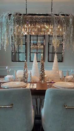 a dining room table decorated for christmas with silver and white decorations on the top, and hanging chandelier above it