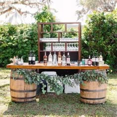 an outdoor bar set up with wine bottles and glasses on top of wooden barrels, surrounded by greenery