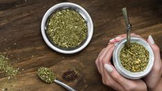 two hands holding small white bowls filled with green herbs and spoons on a wooden table