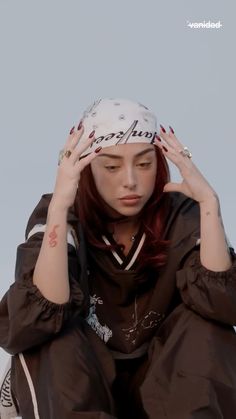 a woman with red hair is sitting down and holding her hands to her head while wearing a hat