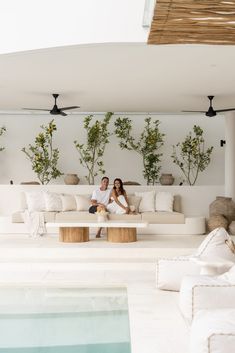 a man and woman sitting on a white couch in front of a wall with potted plants