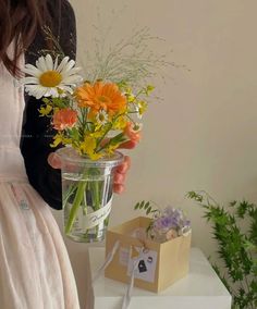 a woman holding a vase filled with flowers on top of a white table next to a plant