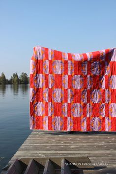 a red and white quilt sitting on top of a wooden dock next to the water